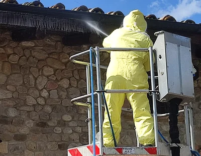 Operatore in tuta protettiva che effettua la disinfestazione di vespe sul tetto usando una piattaforma aerea e motopompa ad alta pressione.